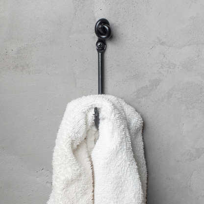 An Urban Forge Knot hand-forged iron single hook in a black finish. The hook is mounted on a gray wall and is holding a white towel.