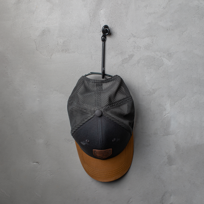 An Urban Forge Knot hand-forged iron single hook in a black finish. The hook is mounted on a gray wall and is holding a ball cap.