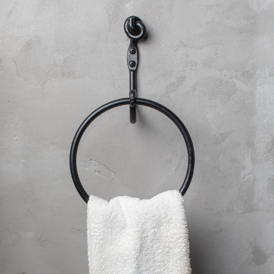 The Urban Forge Knot towel ring in a black finish. The towel ring is mounted on a gray wall and is holding a towel.