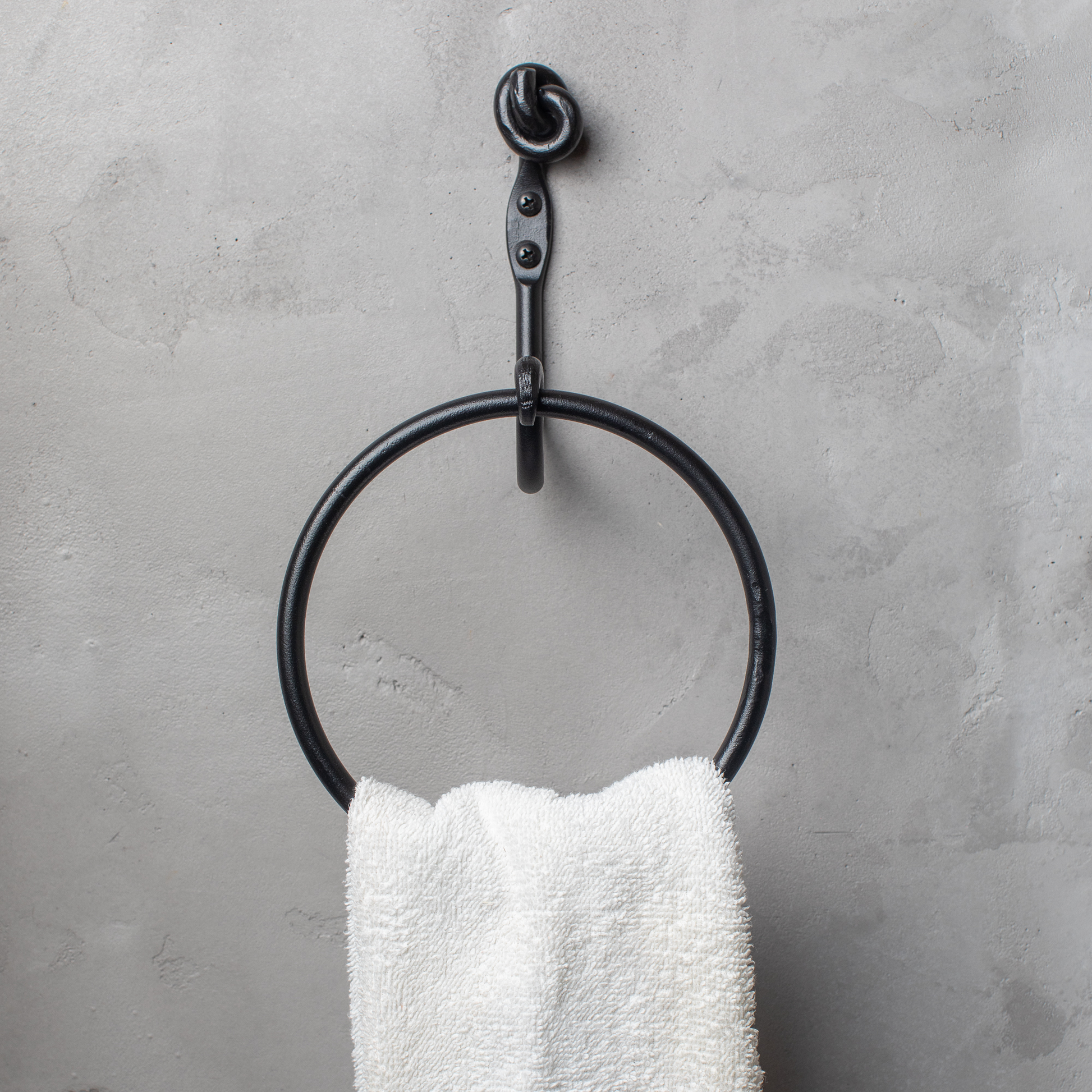 The Urban Forge Knot towel ring in a black finish. The towel ring is mounted on a gray wall and is holding a towel.
