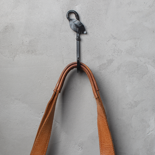 An Urban Forge Leaf hand-forged iron single hook in a black finish. The hook is mounted on a gray wall and is holding a leather bag.