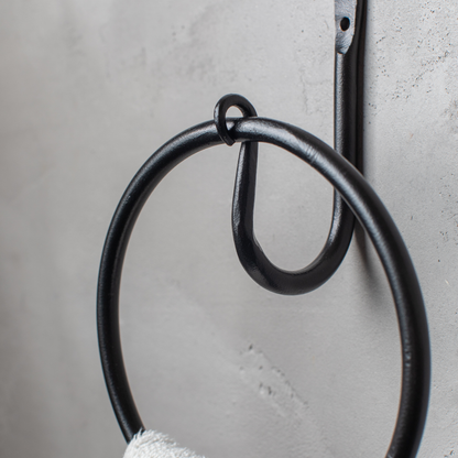 The Urban Forge Leaf towel ring in a black finish. The towel ring is mounted on a gray wall and is holding a towel.