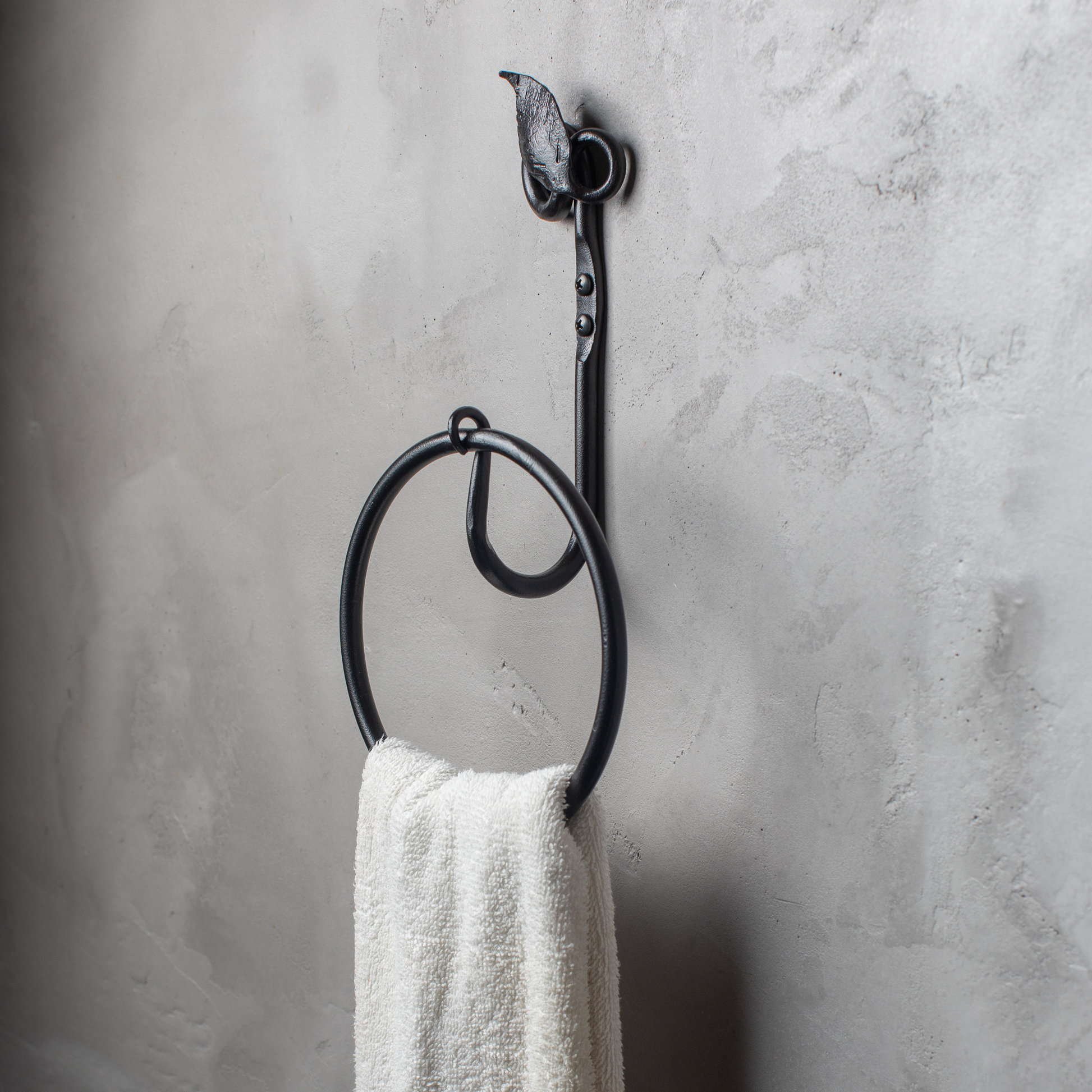 The Urban Forge Leaf towel ring in a black finish. The towel ring is mounted on a gray wall and is holding a towel.