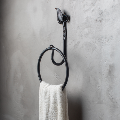 The Urban Forge Leaf towel ring in a black finish. The towel ring is mounted on a gray wall and is holding a towel.