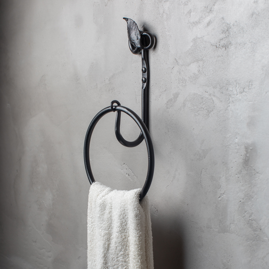 The Urban Forge Leaf towel ring in a black finish. The towel ring is mounted on a gray wall and is holding a towel.