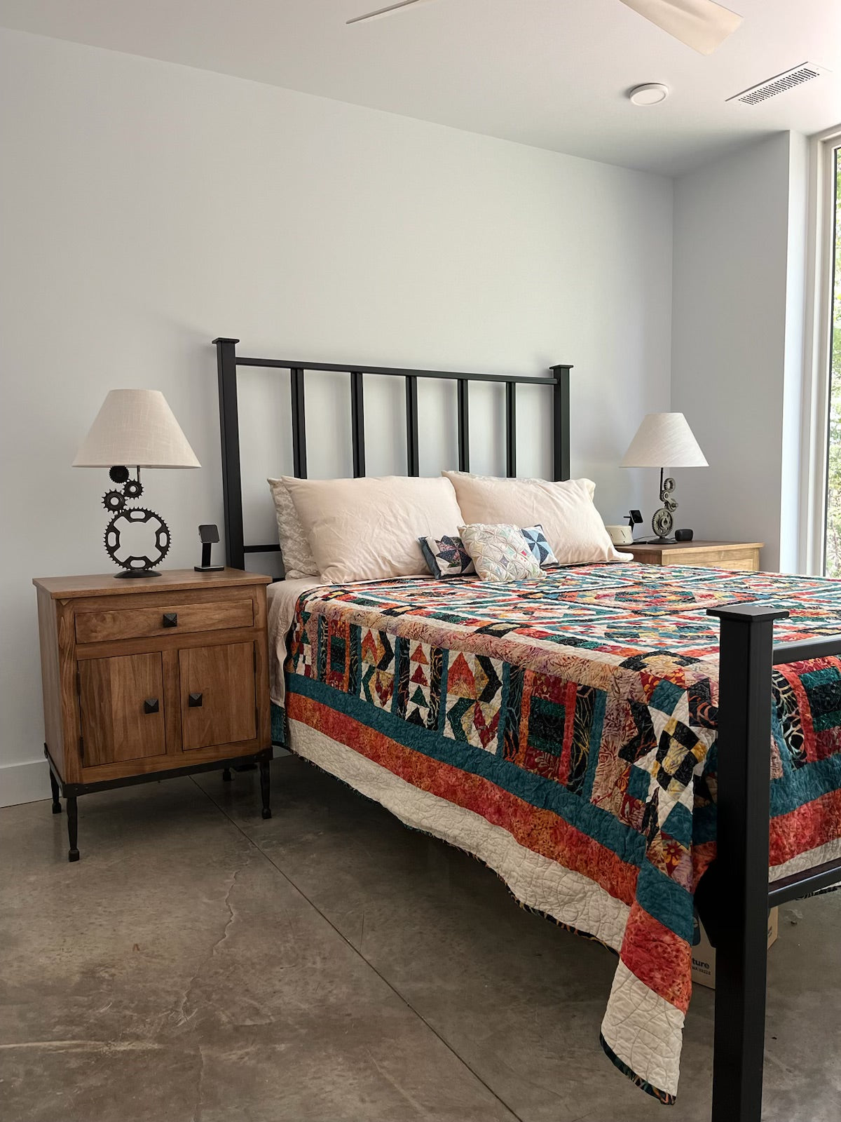 Iron bed with colorful quilt and a wooden and iron side table in a room with a window.