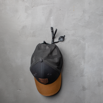 An Urban Forge Sylamore hand-forged iron double hook in a black finish. The hook is mounted on a gray wall and is holding a ball cap.