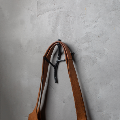 An Urban Forge Sylamore hand-forged iron double hook in a black finish. The hook is mounted on a gray wall and is holding a leather bag.