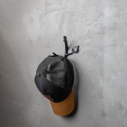 An Urban Forge Sylamore hand-forged iron double hook in a black finish. The hook is mounted on a gray wall and is holding a ball cap.