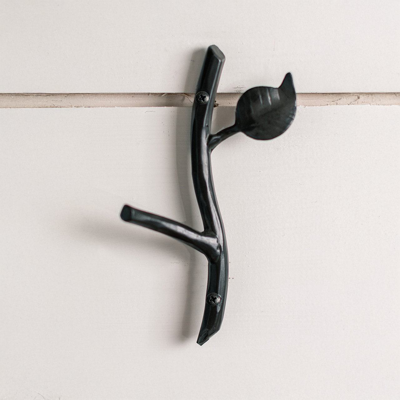 An Urban Forge Sylamore hand-forged iron single hook in a black finish. The hook is mounted on a white shiplap wall.