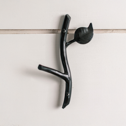 An Urban Forge Sylamore hand-forged iron single hook in a black finish. The hook is mounted on a white shiplap wall.