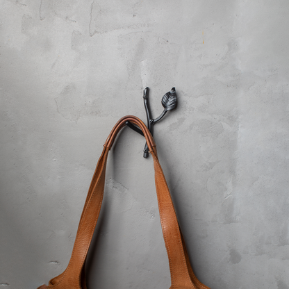 An Urban Forge Sylamore hand-forged iron single hook in a black finish. The hook is mounted on a gray wall and is holding a leather bag.