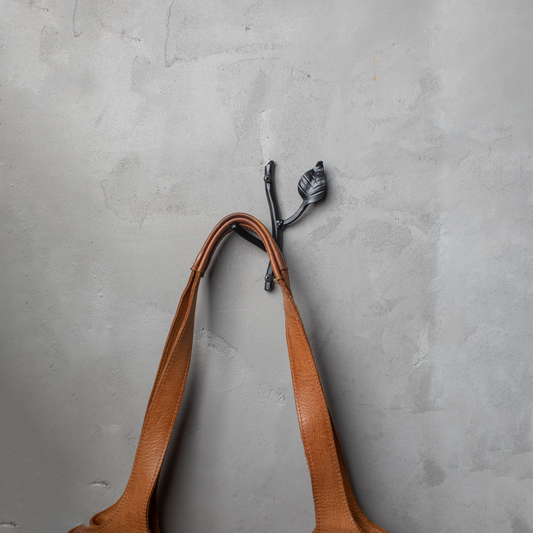 An Urban Forge Sylamore hand-forged iron single hook in a black finish. The hook is mounted on a gray wall and is holding a leather bag.