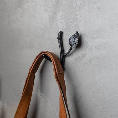 An Urban Forge Sylamore hand-forged iron single hook in a black finish. The hook is mounted on a gray wall and is holding a leather bag.