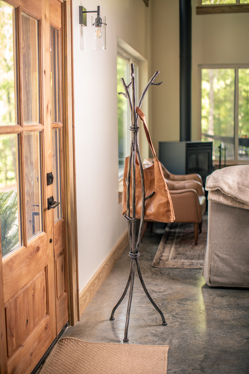 An Urban Forge Sylamore iron standing coat rack in a silver finish. The coat rack is holding a leather bag.