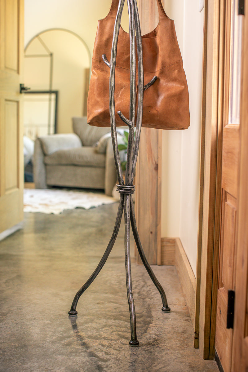 An Urban Forge Sylamore iron standing coat rack in a silver finish. The coat rack is holding a leather bag.