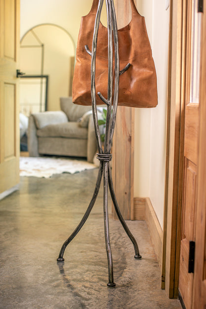 An Urban Forge Sylamore iron standing coat rack in a silver finish. The coat rack is holding a leather bag.